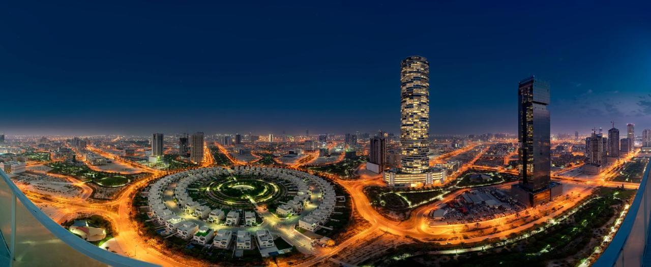 Five Jumeirah Village Dubai Esterno foto The photo showcases a cityscape illuminated at night. In the foreground, there is a circular arrangement of buildings or houses, creating a visually striking pattern. To the right, a tall, modern skyscraper stands out, featuring a sleek design with n