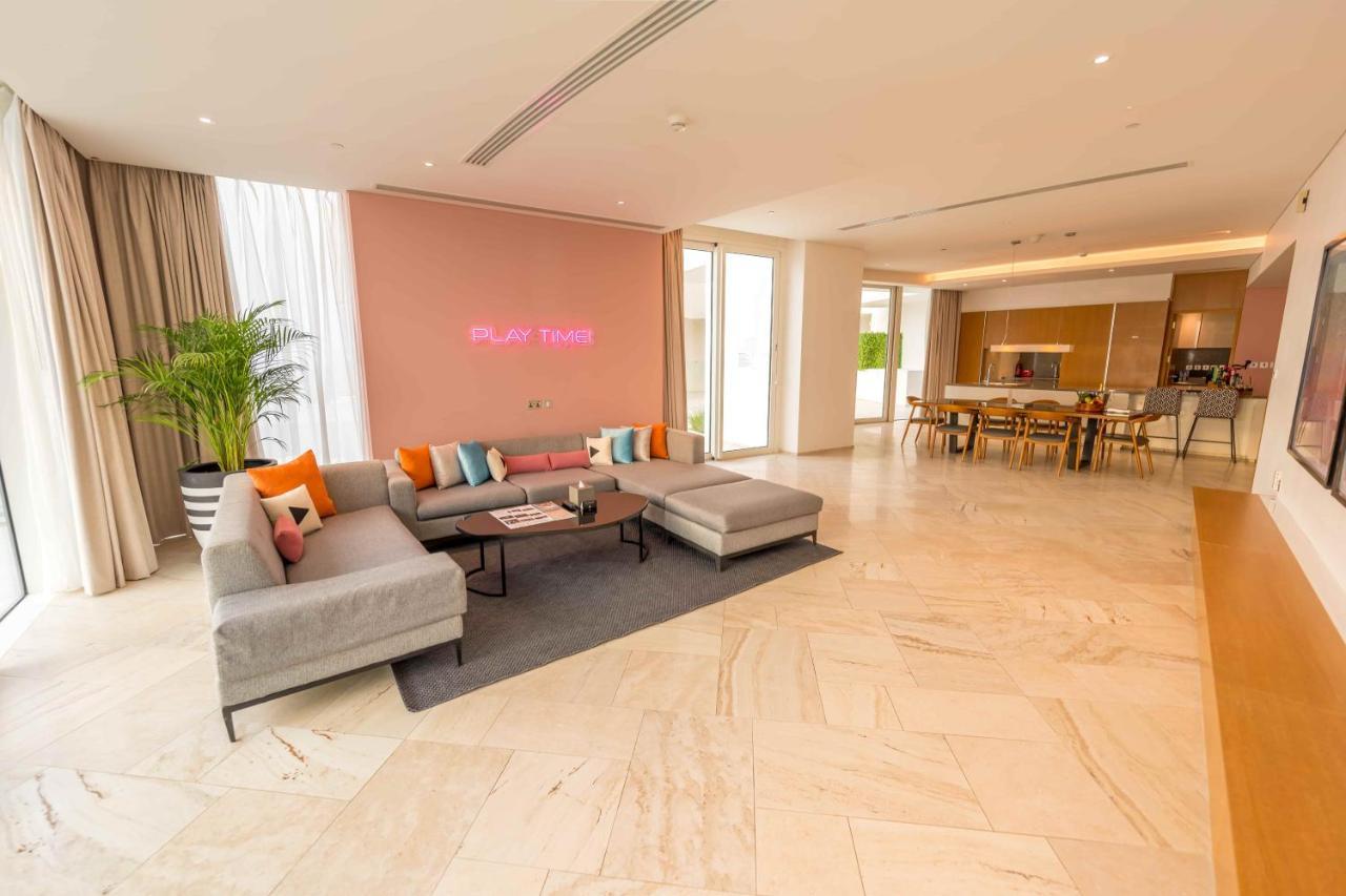 Five Jumeirah Village Dubai Esterno foto The photo shows a spacious and modern living area. There is a large, light-colored sectional sofa with colorful cushions, positioned on a dark rug. The wall behind the sofa is painted in a soft pink shade, and there is a subtle decorative element or 
