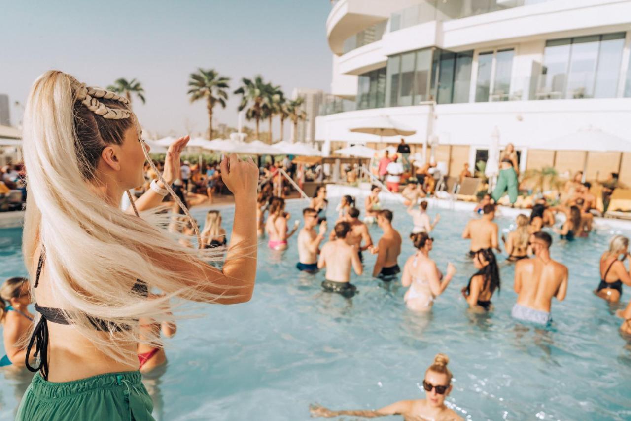 Five Jumeirah Village Dubai Esterno foto The photo features a lively pool party scene. In the foreground, a woman with long, blonde hair is seen raising her arm as if enjoying the moment. The pool is filled with many people, some swimming and others socializing. The atmosphere appears festi