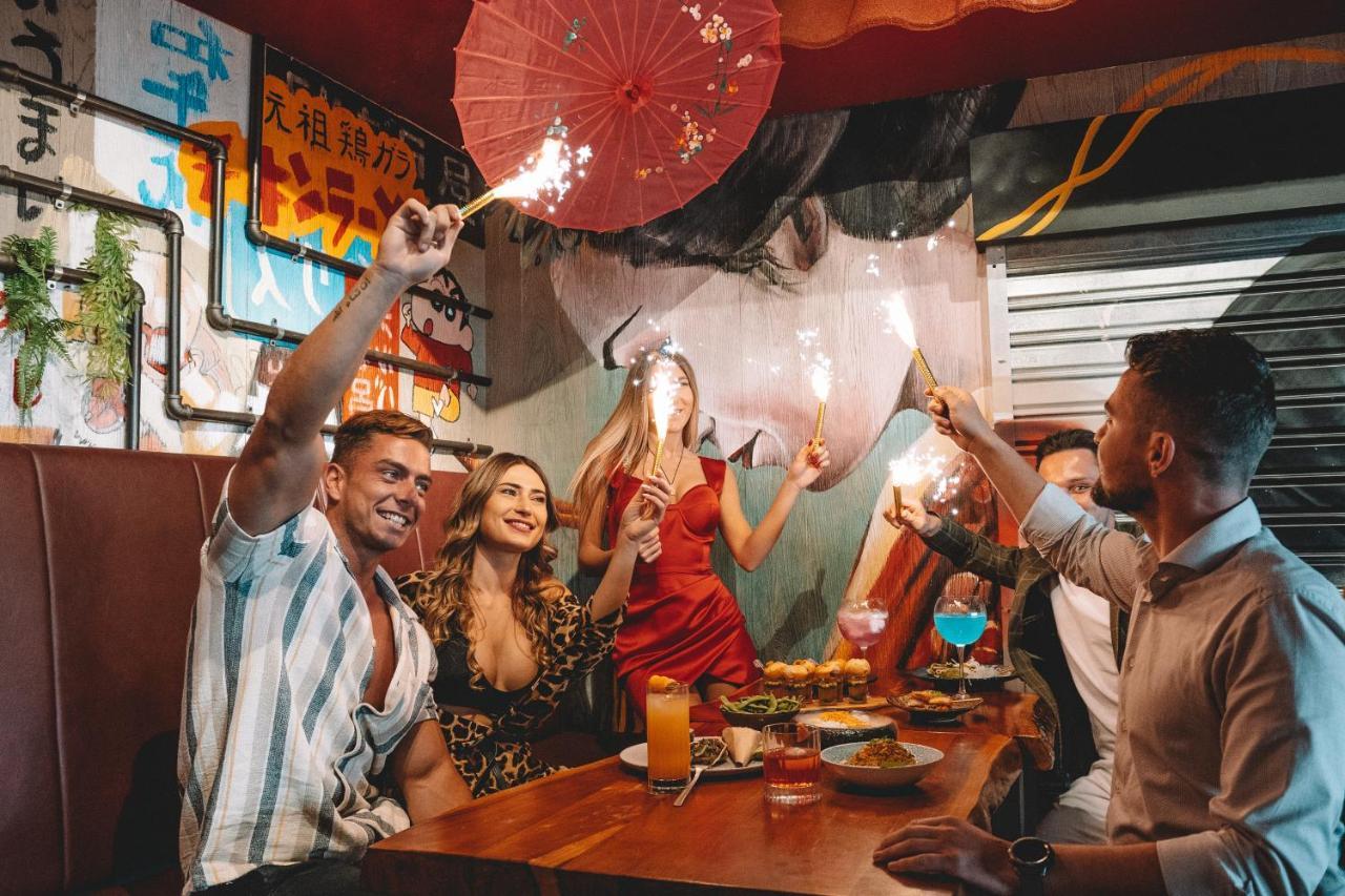 Five Jumeirah Village Dubai Esterno foto The photo depicts a lively gathering at a dining table where several people are celebrating. There are four individuals: two men and two women. One of the women is wearing a red dress, while the other is dressed in a patterned outfit. They are holdin