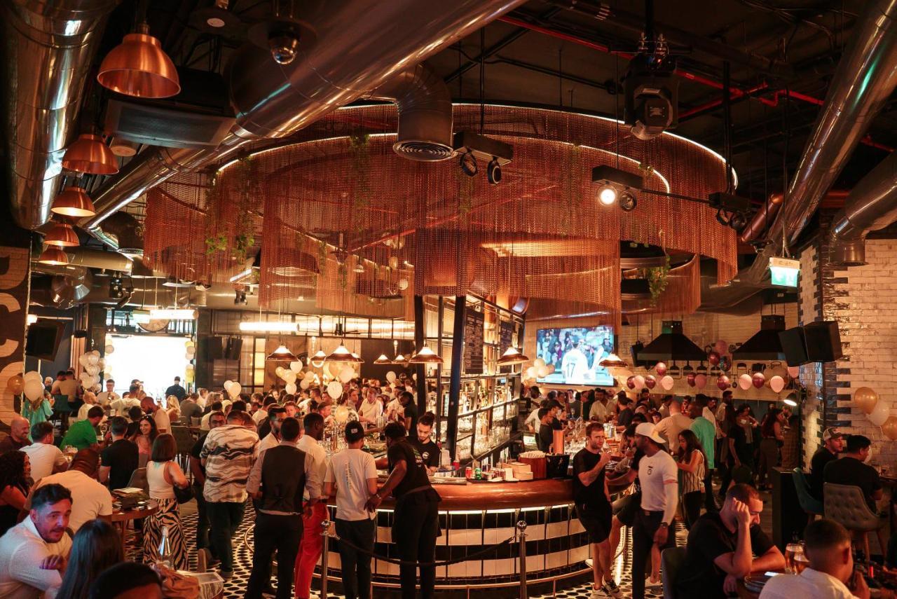 Five Jumeirah Village Dubai Esterno foto The photo depicts a lively bar or restaurant scene filled with people socializing. The interior design features a modern aesthetic with a mix of industrial elements, such as exposed pipes and metallic accents. A large, circular bar is the focal point