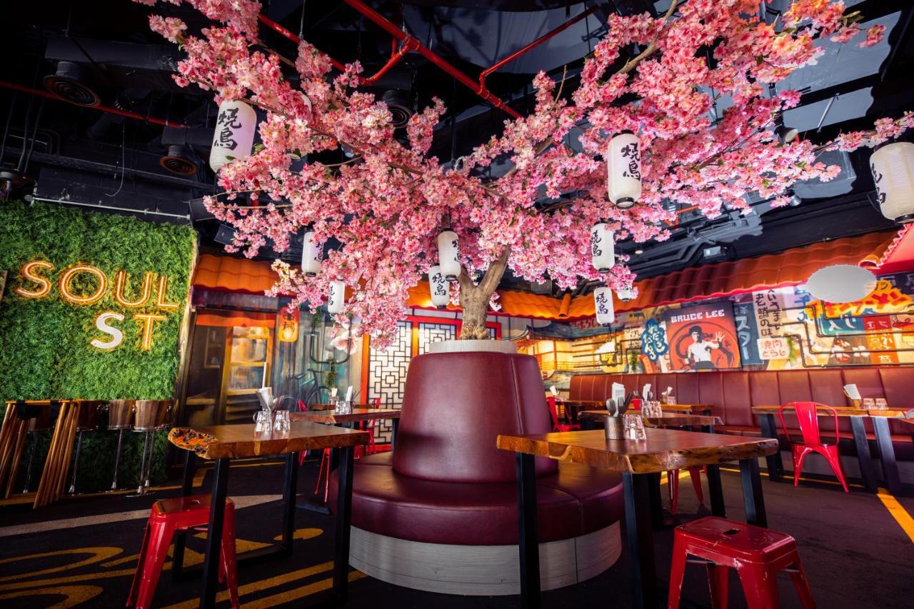 Five Jumeirah Village Dubai Esterno foto The photo shows a vibrant and modern restaurant interior. At the center, there is a large cherry blossom tree adorned with pink flowers and hanging lanterns, creating a lively atmosphere. Surrounding the tree is a circular booth with maroon upholster