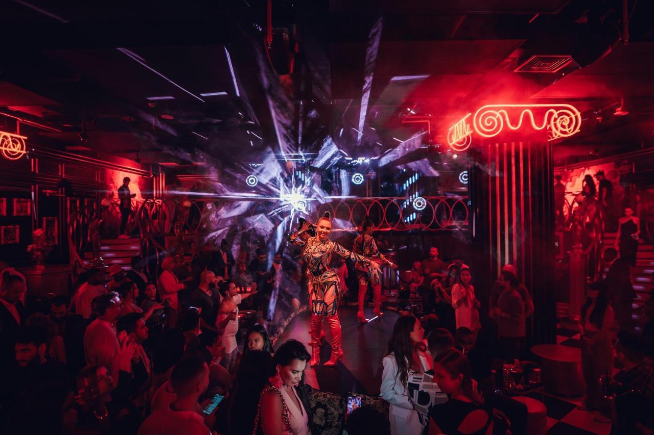 Five Jumeirah Village Dubai Esterno foto The photo depicts a vibrant nightlife scene, likely from a nightclub or performance venue. The setting is dimly lit with striking red lights and dynamic visual effects, creating an electrifying atmosphere. In the center, a performer dressed in a flas