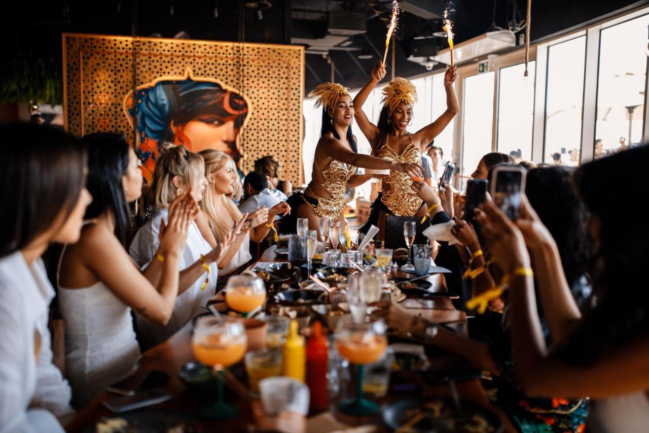 Five Jumeirah Village Dubai Esterno foto The photo depicts a lively restaurant scene where two performers are entertaining a group of seated diners. The performers, dressed in colorful and elaborate outfits, are holding sparklers, adding a festive atmosphere to the occasion. The diners, mos