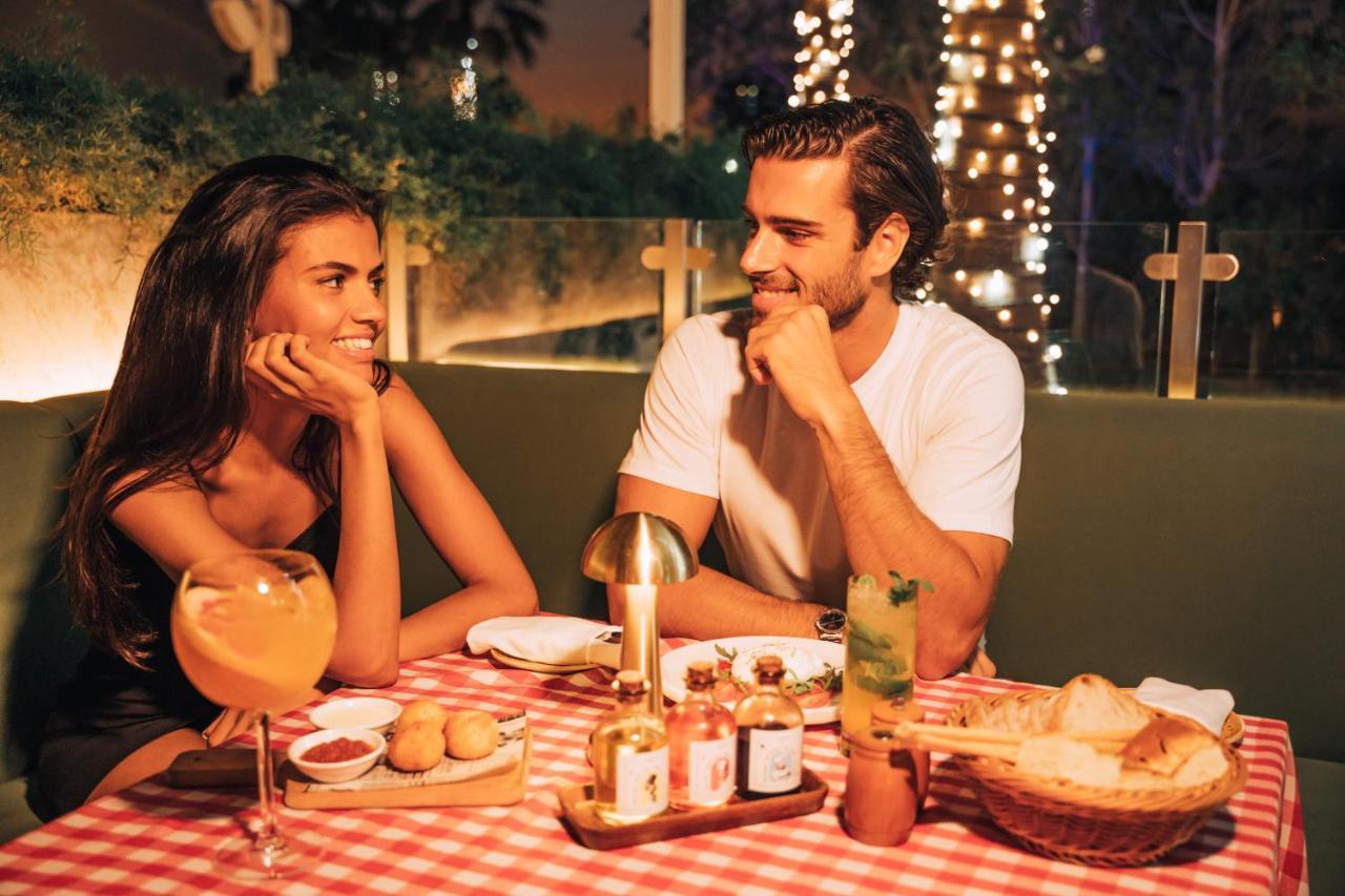 Five Jumeirah Village Dubai Esterno foto The photo shows a romantic dining scene featuring a couple at a table. The setting is illuminated with soft lights, creating a warm atmosphere. The table is adorned with a red and white checkered tablecloth, and various dishes are presented, includin
