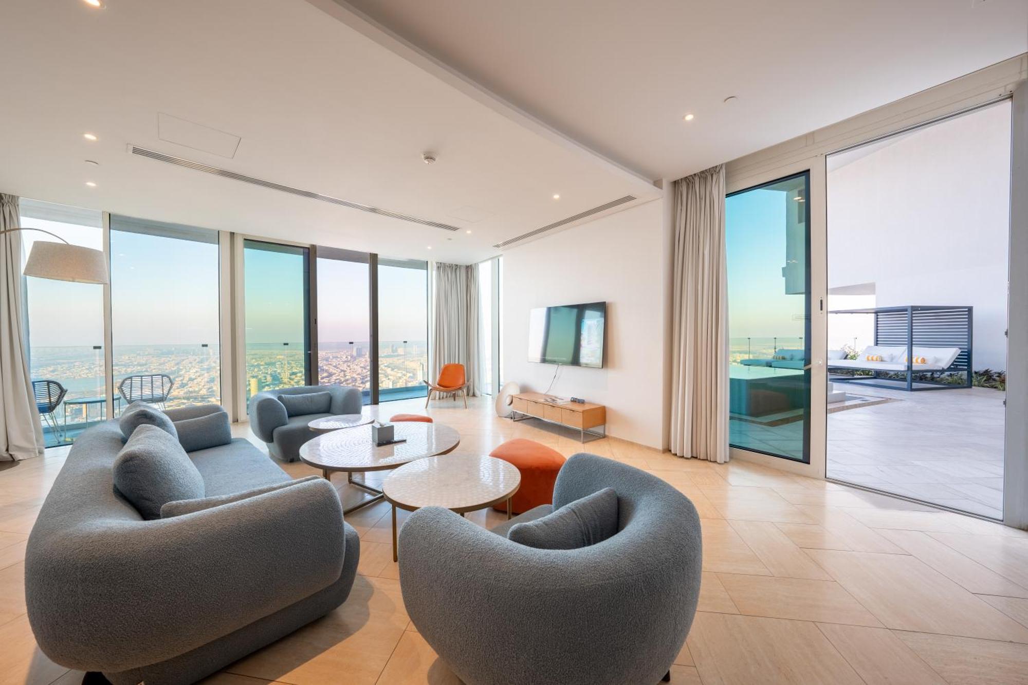 Five Jumeirah Village Dubai Esterno foto The photo shows a modern and stylish living room area with large windows that provide a scenic view. There are several rounded, gray sofas arranged around a couple of round coffee tables. A sleek television is mounted on the wall across from the seat