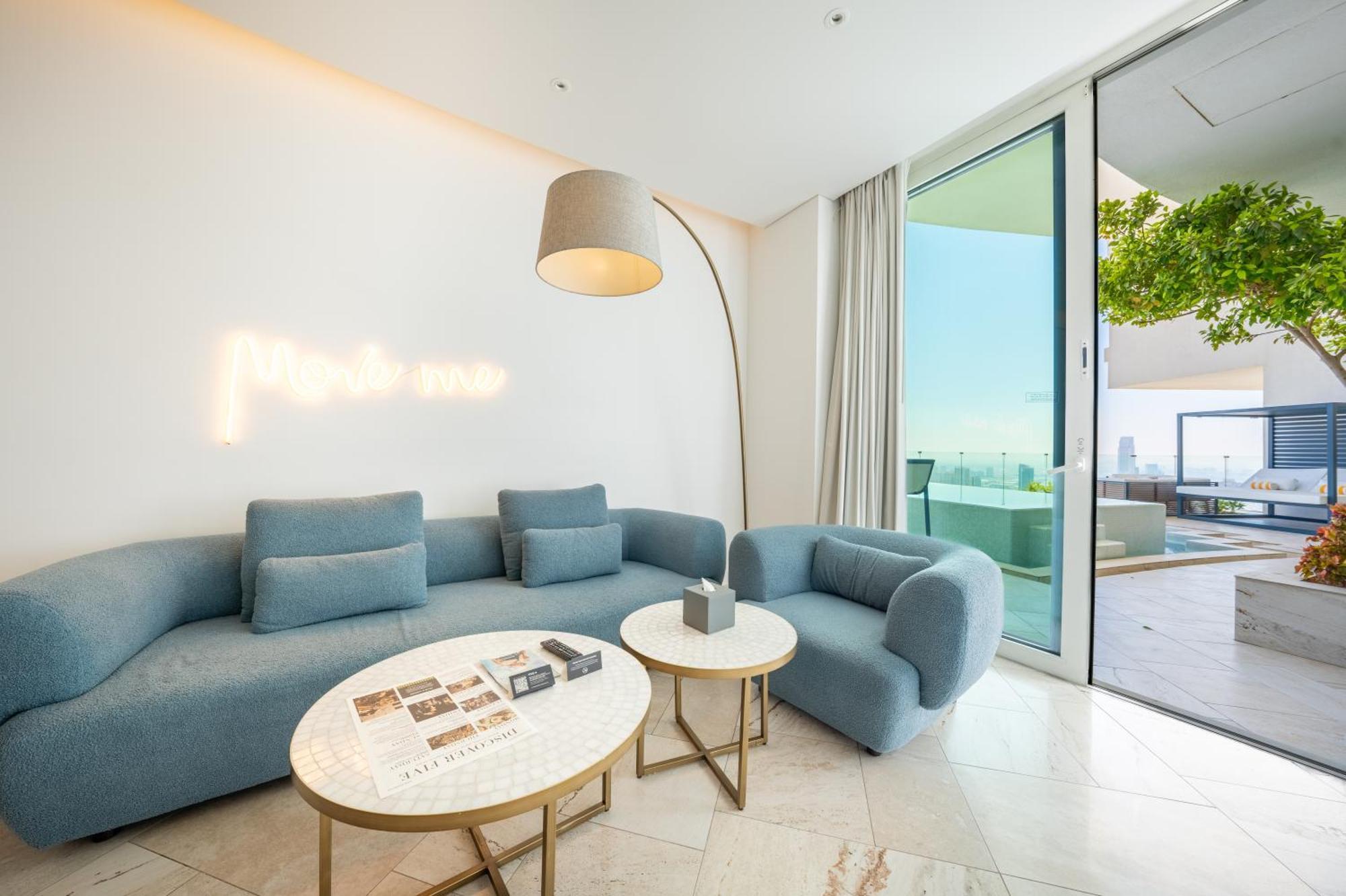 Five Jumeirah Village Dubai Esterno foto The photo shows a modern and stylish living space. It features a light blue sectional sofa and a matching armchair, creating a cozy seating area. Two round marble coffee tables are placed in front, one of which has some magazines or books on it. A ta