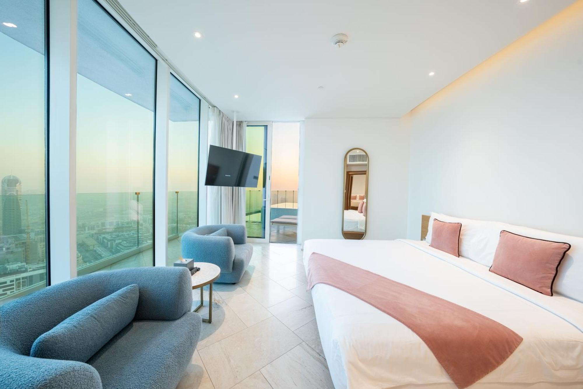 Five Jumeirah Village Dubai Esterno foto The photo depicts a modern hotel room featuring large windows that provide a view of the outside. The room includes a spacious bed with a pink accent blanket, a cozy blue sofa, a small round table, and a wall-mounted television. There is also a mirro