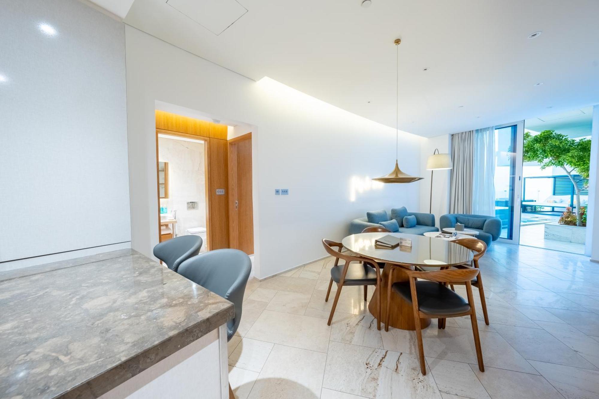 Five Jumeirah Village Dubai Esterno foto The photo shows a modern living space that combines a dining area and a lounging area. On the left, there is a kitchen counter with a marble surface and several gray chairs around it. In the center, there is a round dining table made of wood with sev