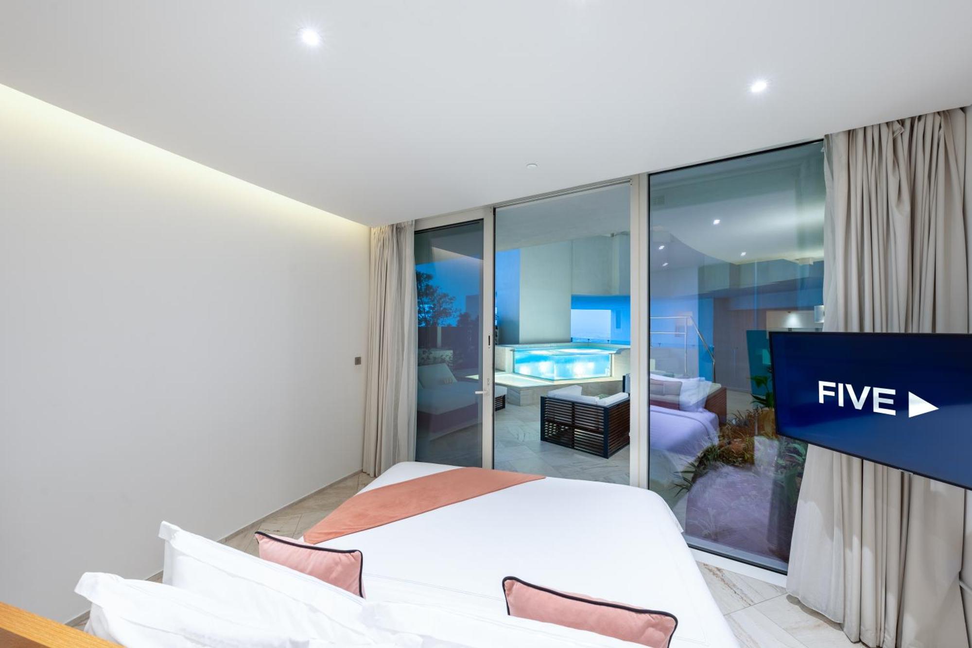 Five Jumeirah Village Dubai Esterno foto The photo shows a modern, stylish bedroom featuring a large bed with light-colored bedding and decorative pillows. There are large glass windows that open up to a view of an outdoor area, likely overlooking a pool or patio. The room has neutral-color