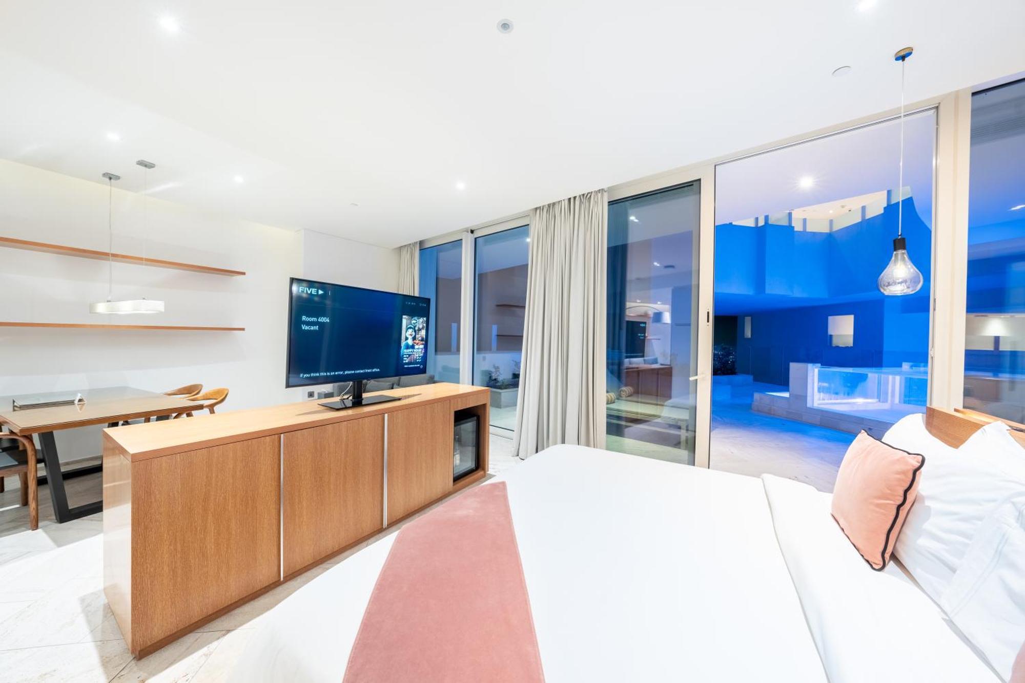 Five Jumeirah Village Dubai Esterno foto The photo depicts a modern hotel room with a clean and minimalist design. A large bed is shown, topped with white linens and decorative pillows. Opposite the bed, there is a wooden TV stand with a flat-screen television. The room features large windo