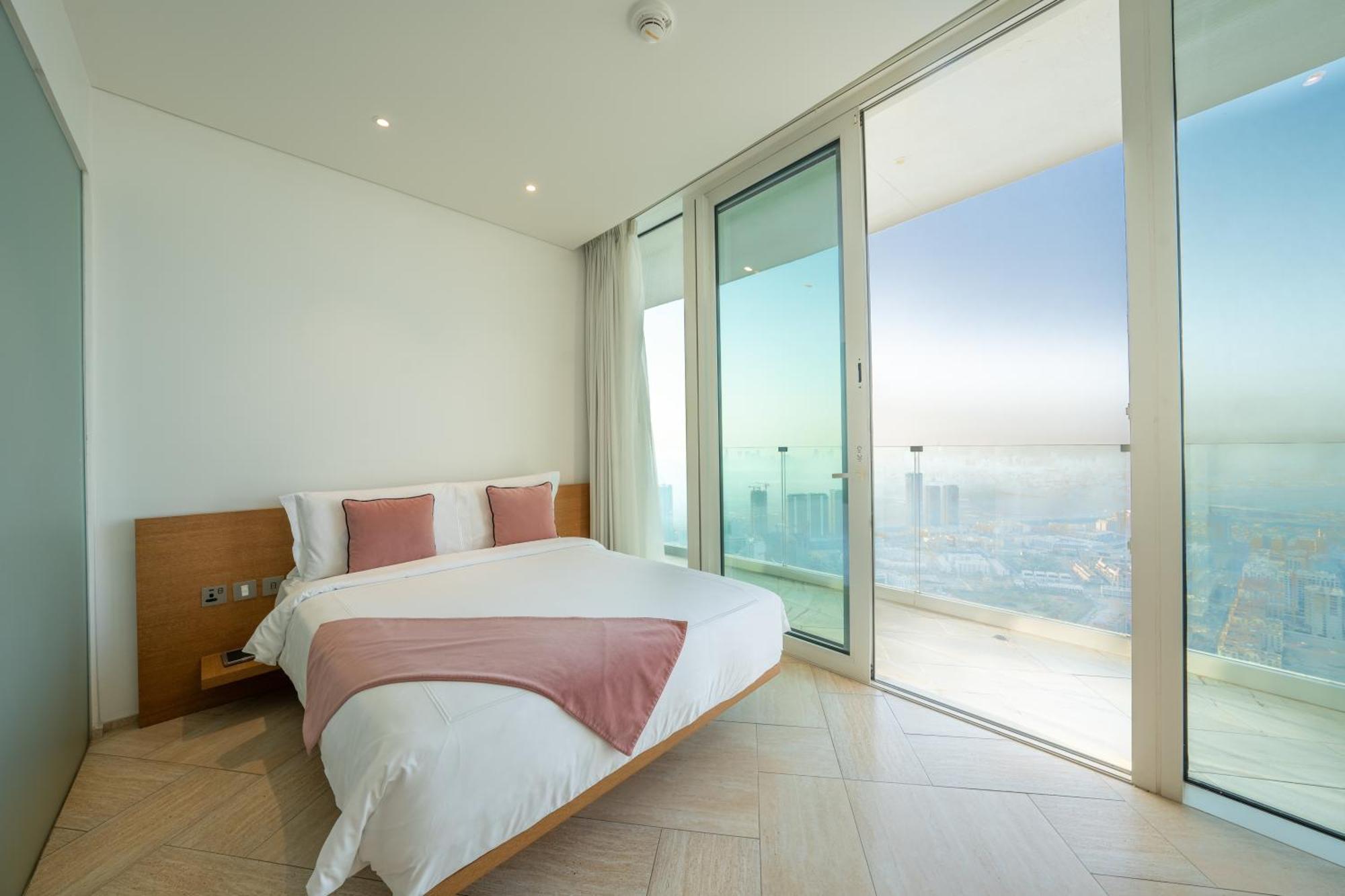 Five Jumeirah Village Dubai Esterno foto The photo shows a modern bedroom with a large bed featuring white bedding and accent pillows in a soft pink color. The room has floor-to-ceiling glass doors that lead out to a balcony, providing a panoramic view of the outside. The walls are painted 