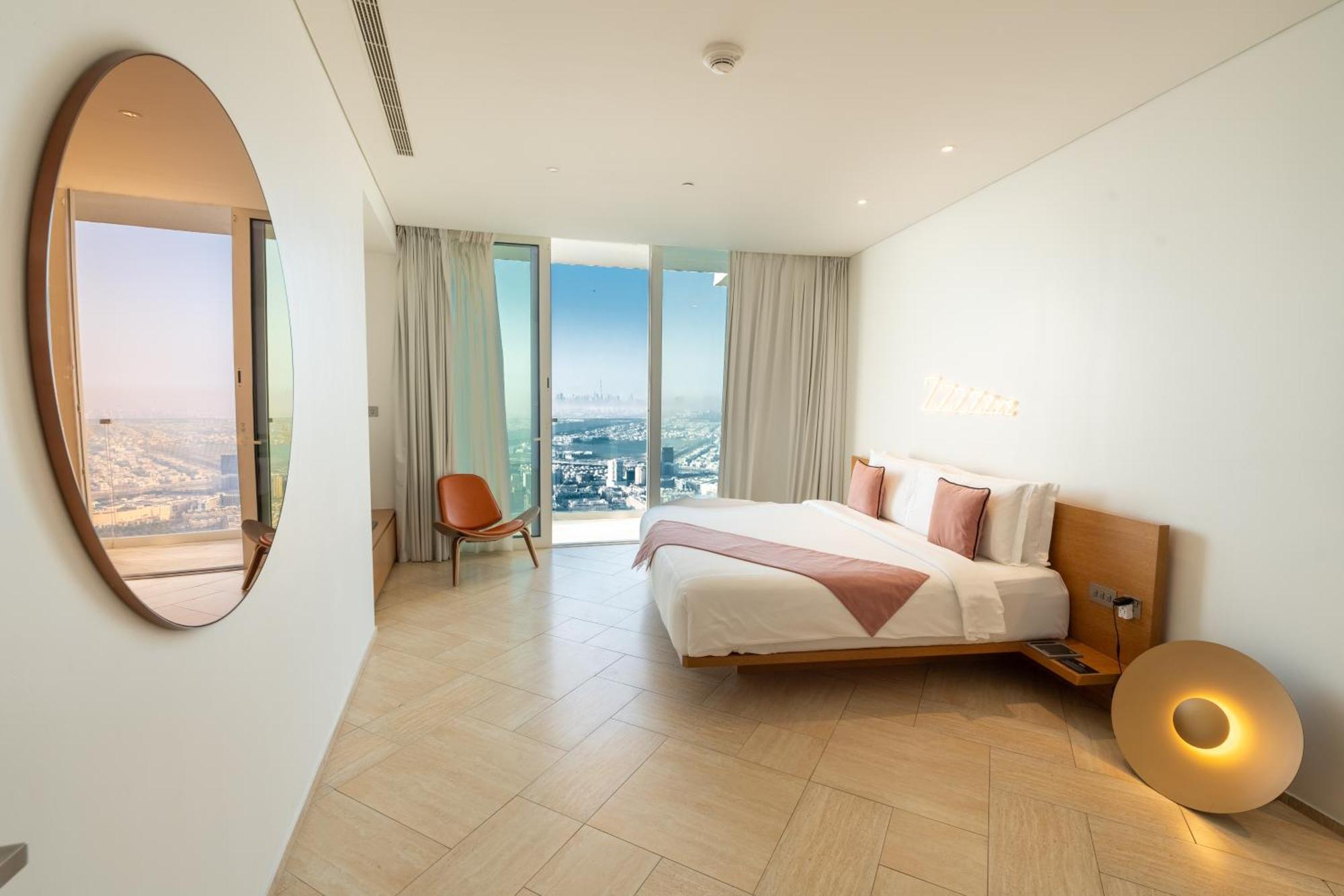 Five Jumeirah Village Dubai Esterno foto The photo shows a modern bedroom with a minimalist design. There is a large bed with white bedding and pink accents, positioned against a wall. To the side, there is a round mirror and a stylish chair. The room features large windows dressed with lig