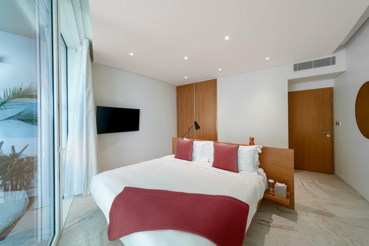 Five Jumeirah Village Dubai Esterno foto The photo shows a modern and stylish bedroom. It features a bed with white bedding and two red decorative pillows. There is a mounted flat-screen TV on the wall opposite the bed. The room has large glass windows that allow natural light to enter, and