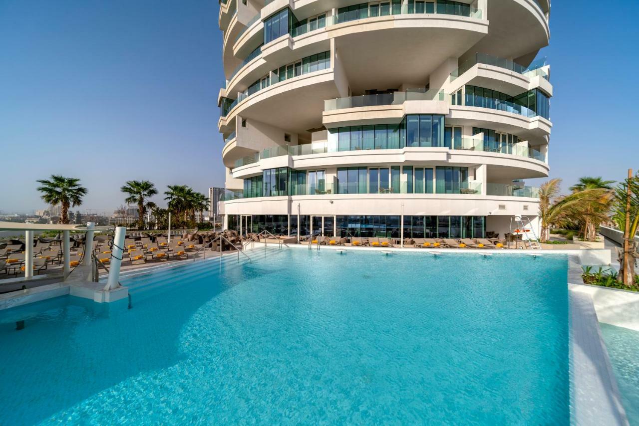 Five Jumeirah Village Dubai Esterno foto The photo depicts a modern building with a distinctive architectural design, featuring multiple curved balconies. In front of the building, there is a large, inviting swimming pool with clear, turquoise water. Surrounding the pool are loungers for re