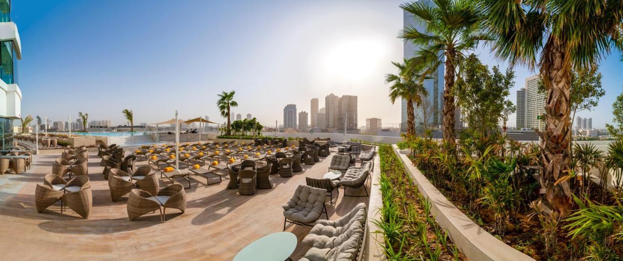 Five Jumeirah Village Dubai Esterno foto The photo showcases a luxurious outdoor seating area, likely part of a resort or hotel. In the foreground, there are well-arranged lounge chairs and seating options, some with cushions, placed on a tiled surface. The area is bordered by lush green pl